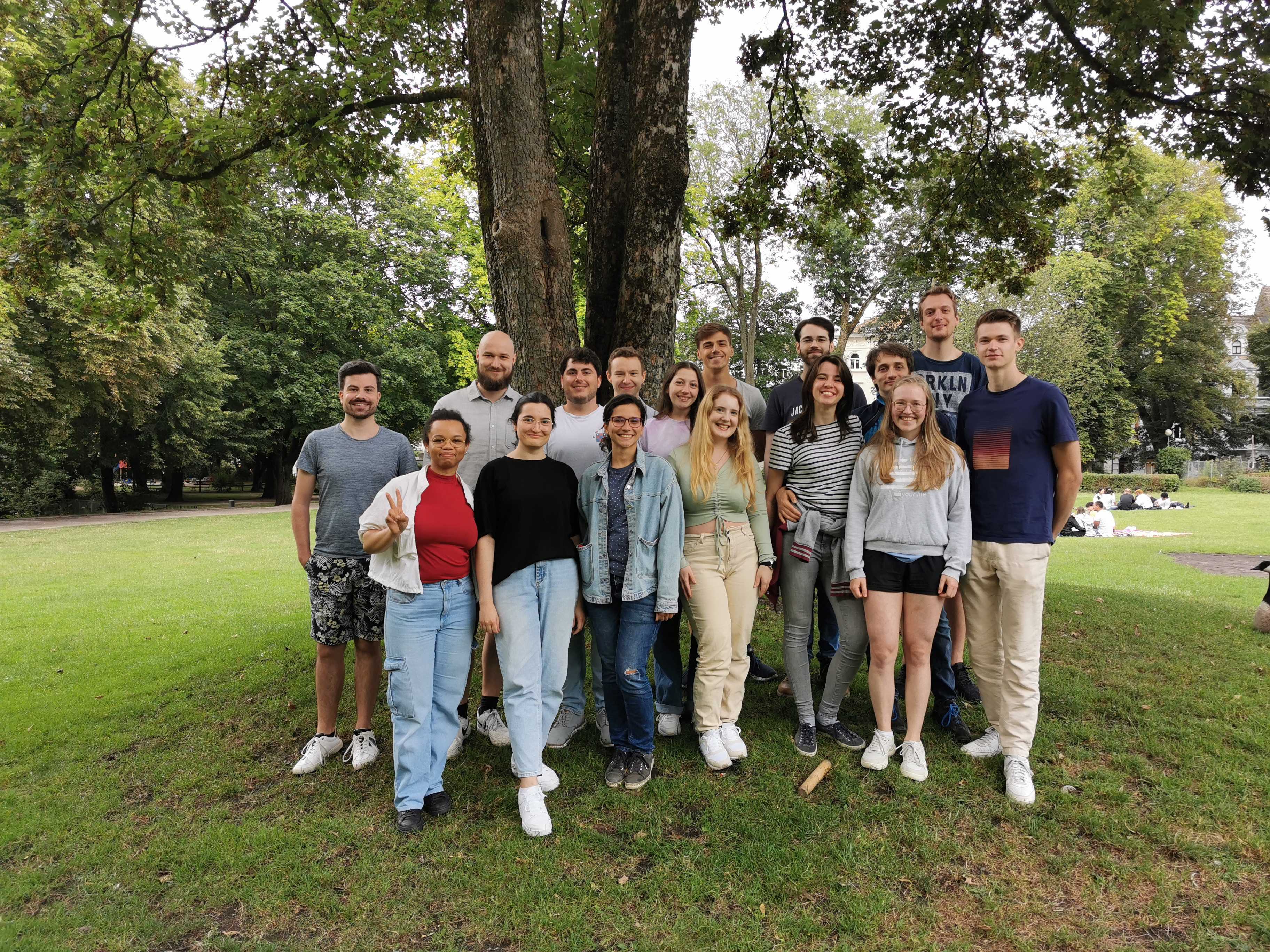 Sechszehn junge Menschen stehen in einem Park vor einem Baum und lächeln in die Kamera